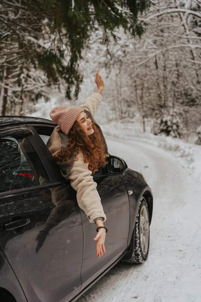 Woman enjoying a winter drive through a snowy forest, leaning out of a car window, smiling joyfully.