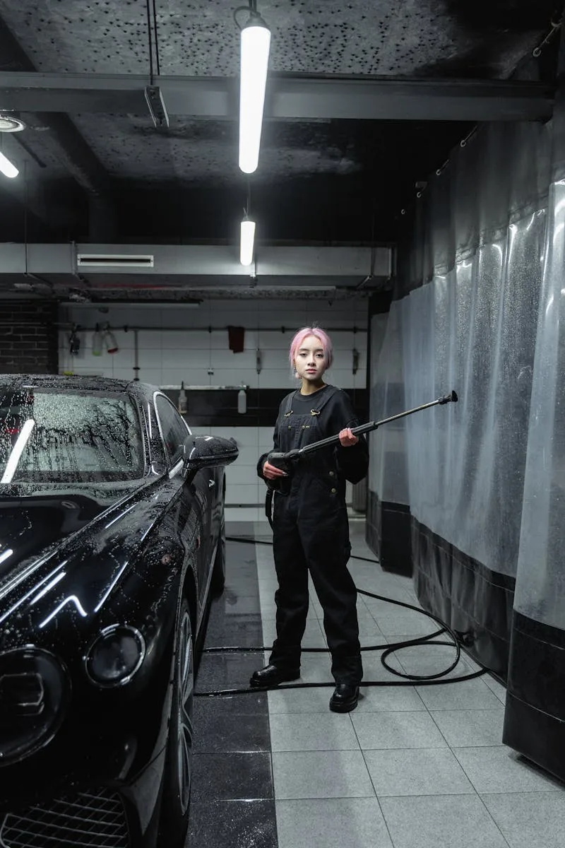 Woman in uniform using power spray at indoor car wash, detailing black luxury car.